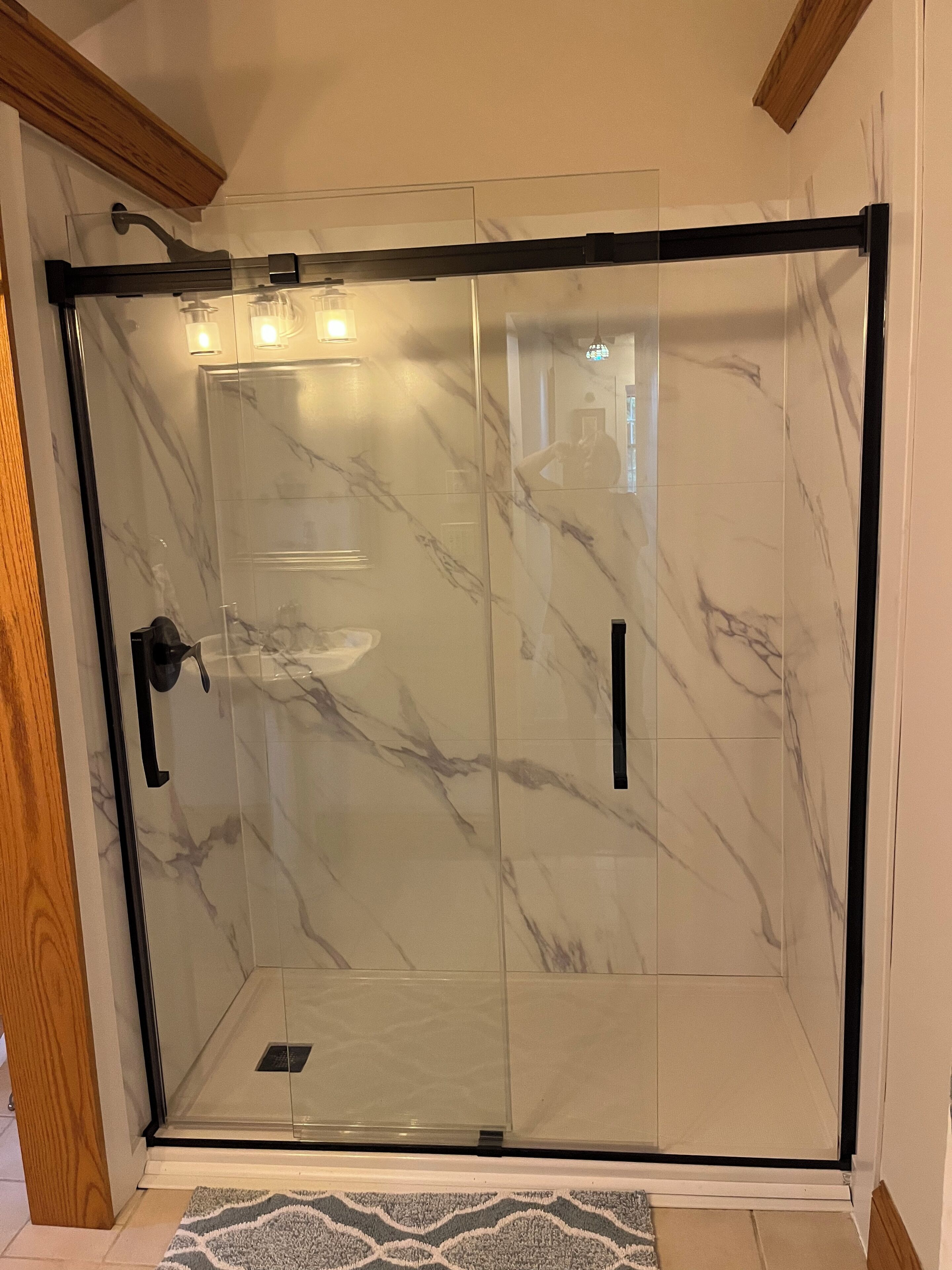 A glass-enclosed shower with a black frame, marble tiles, and a wooden ceiling beam.