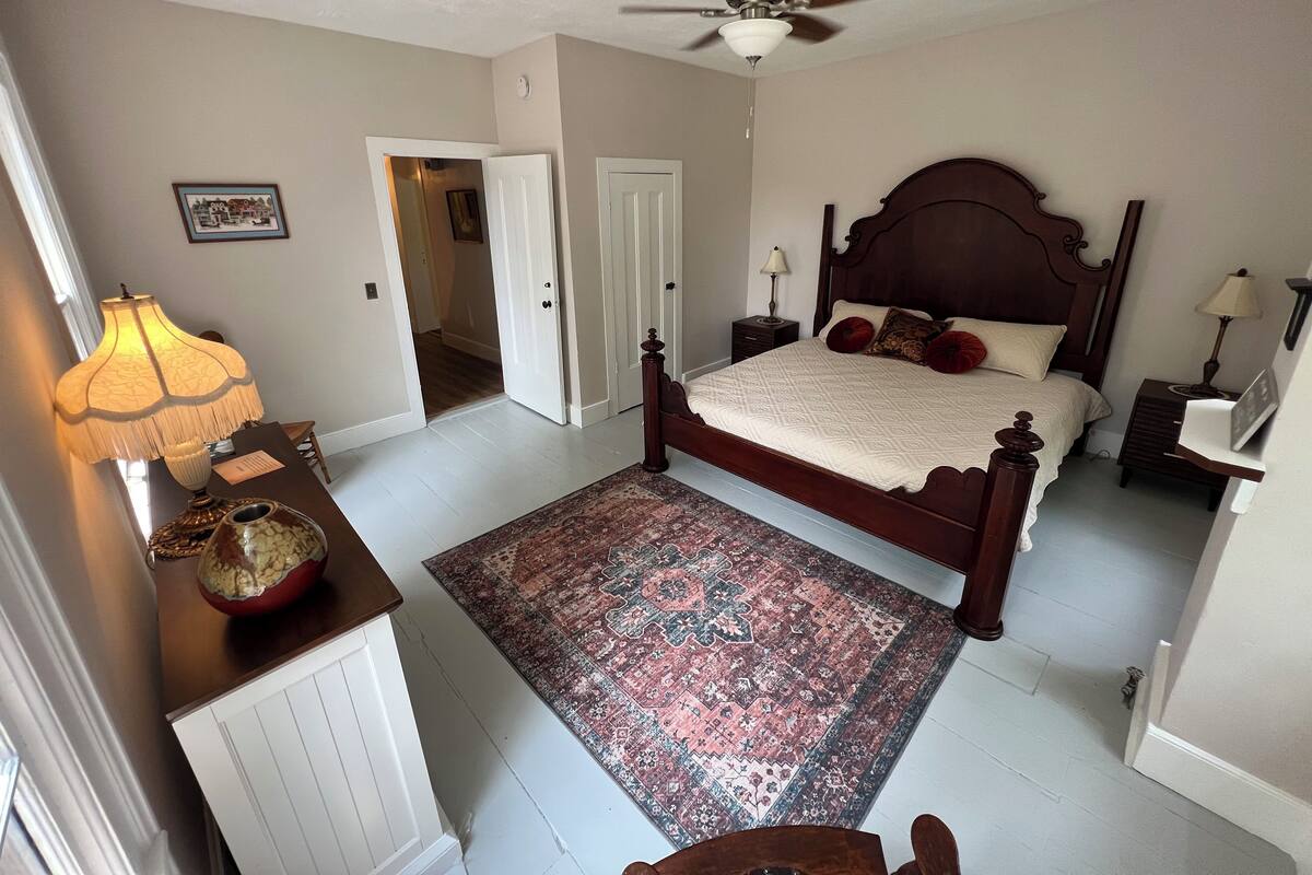 A bedroom with a wooden bed, a ceiling fan, a nightstand with a lamp, a bedside table with a lamp, and a patterned rug.