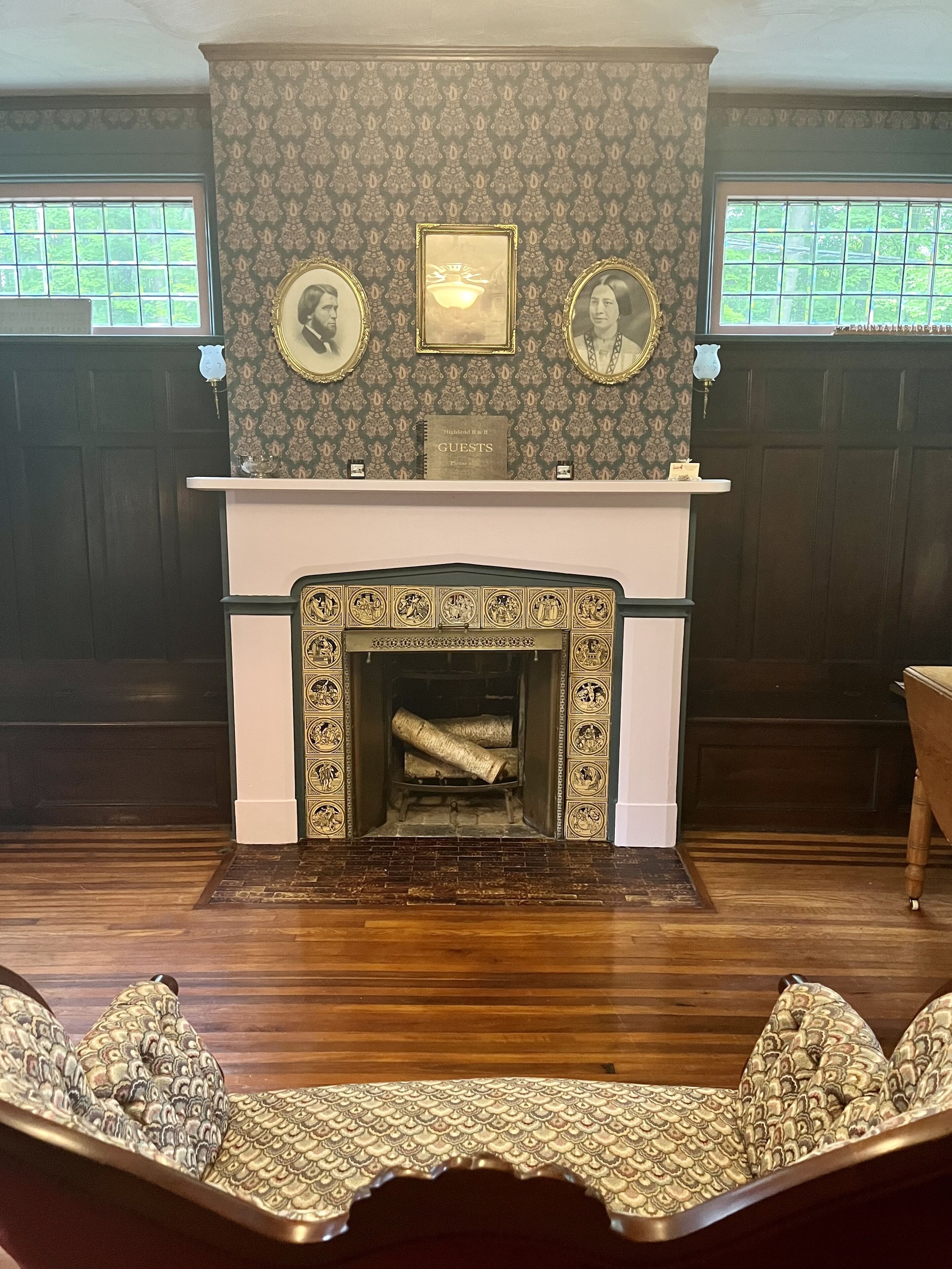 A traditional fireplace with ornate designs, flanked by patterned wallpaper and framed portraits.