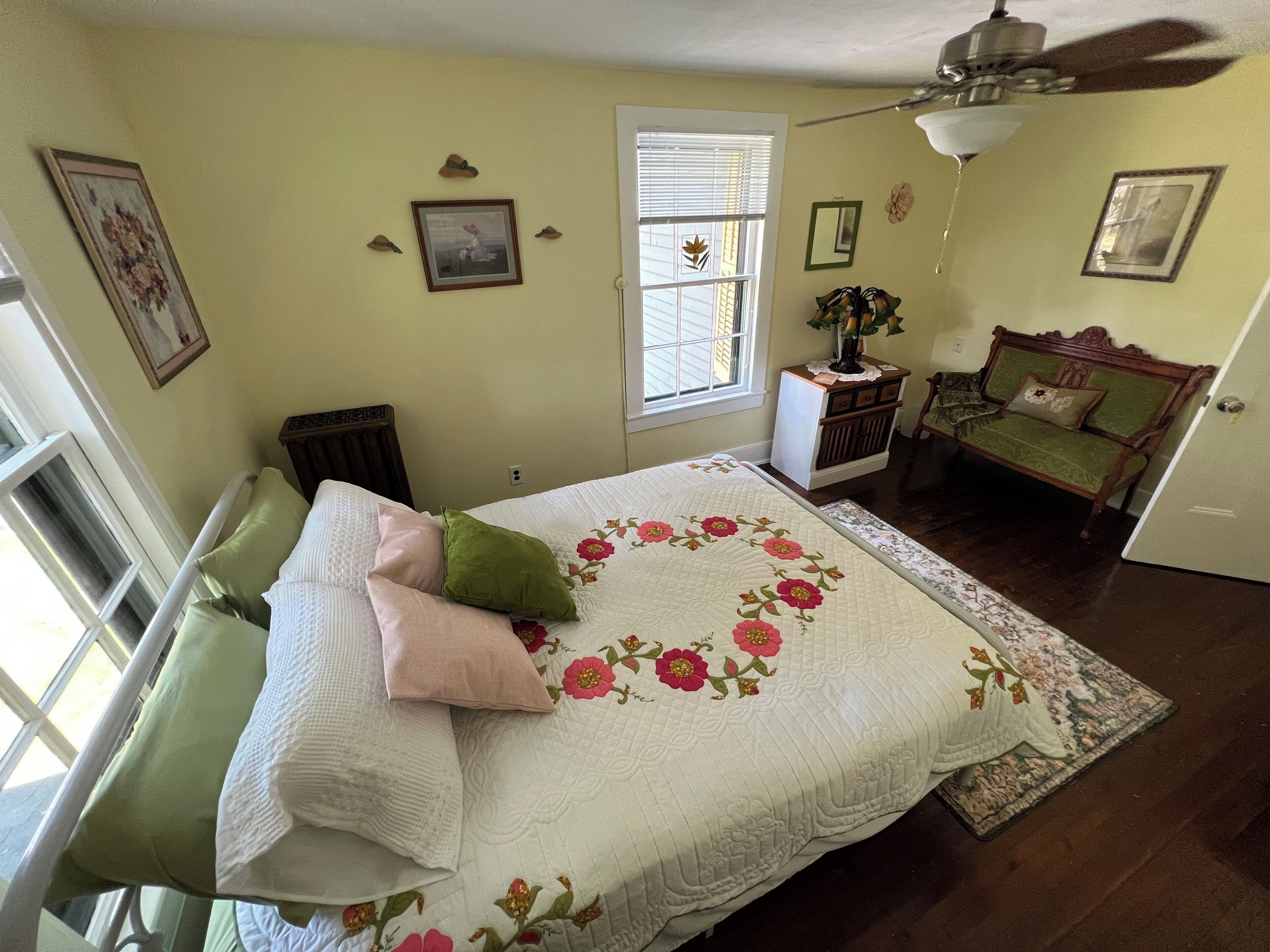Ein Schlafzimmer mit Bett, Nachttisch, Stuhl, Fenster mit Jalousien und Deckenventilator.