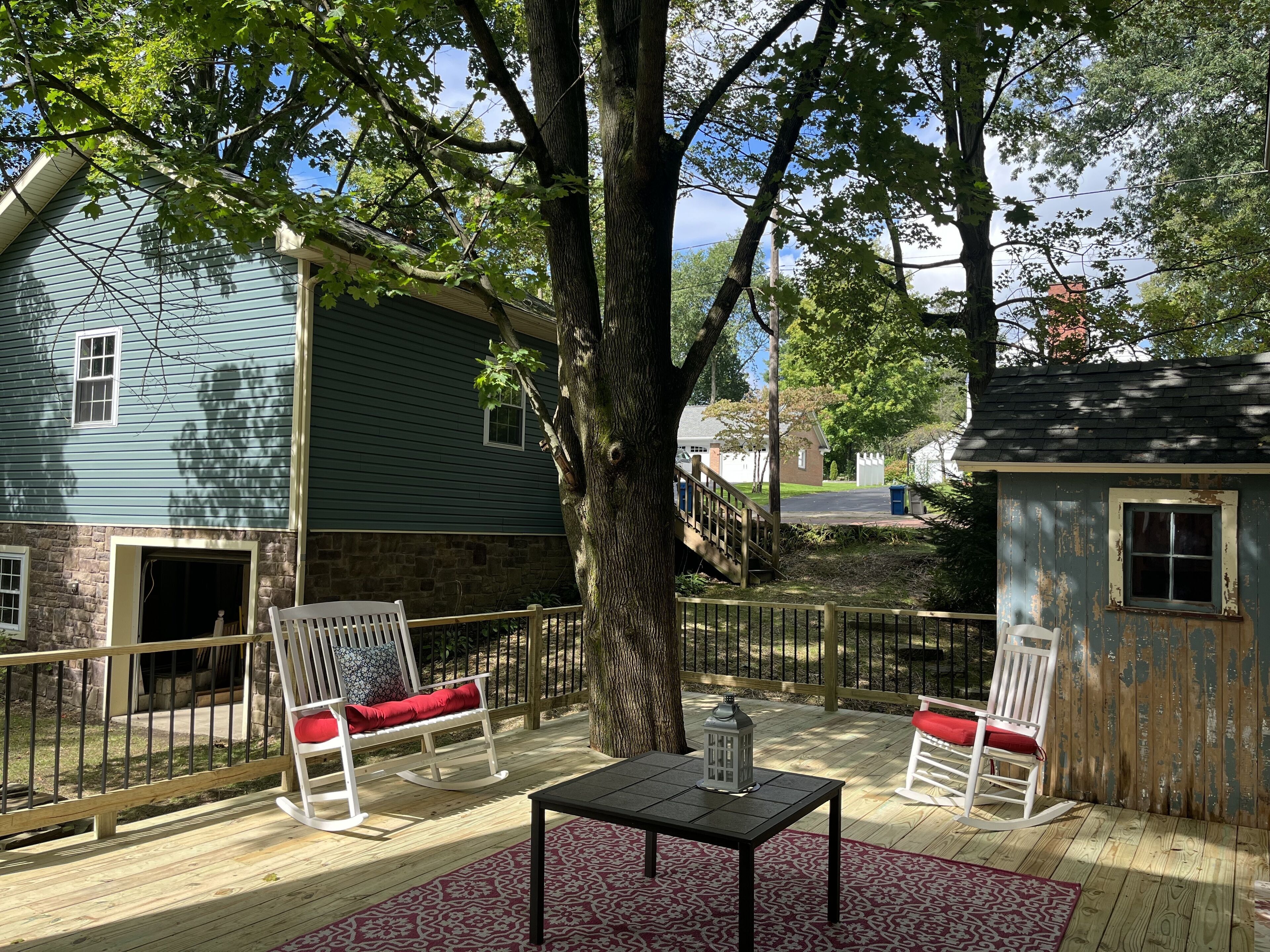 A wooden deck with rocking chairs and a table, surrounded by trees and a house.
