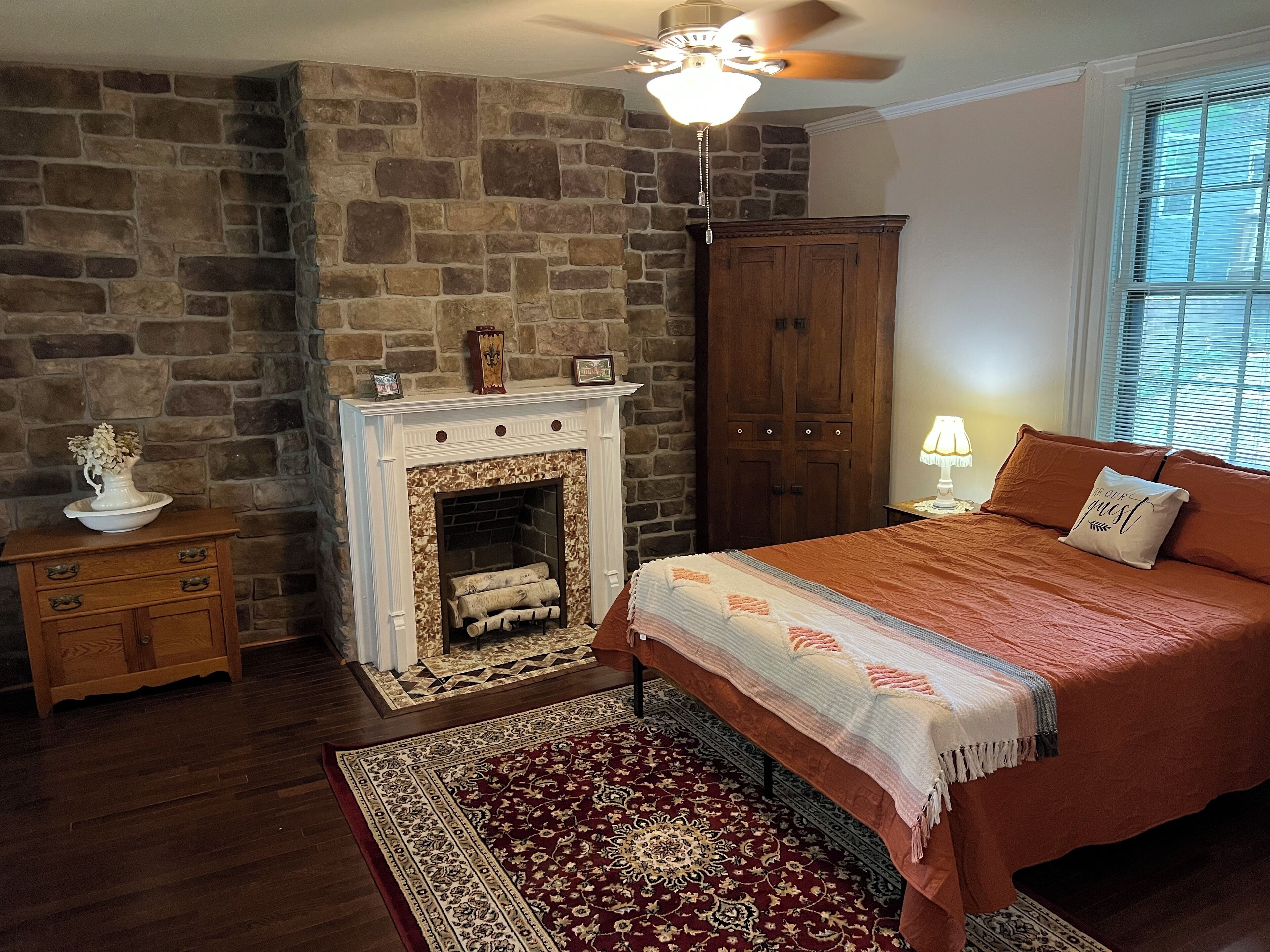 Un dormitorio con chimenea de piedra, una cama con colcha roja, un aparador de madera y una mesita de noche con lámpara.
