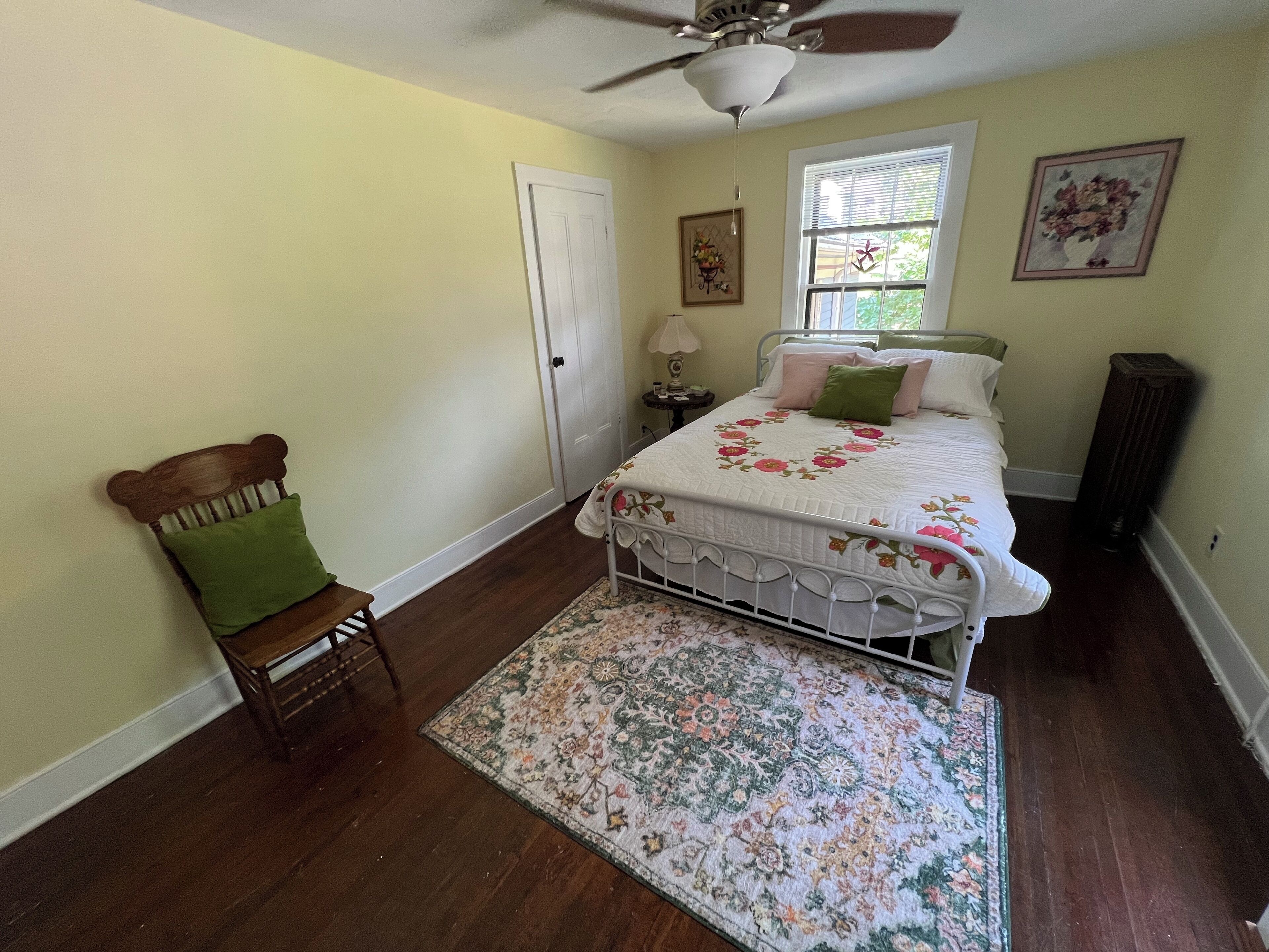 A bedroom with a bed, a chair, a rug, a ceiling fan, and a window.
