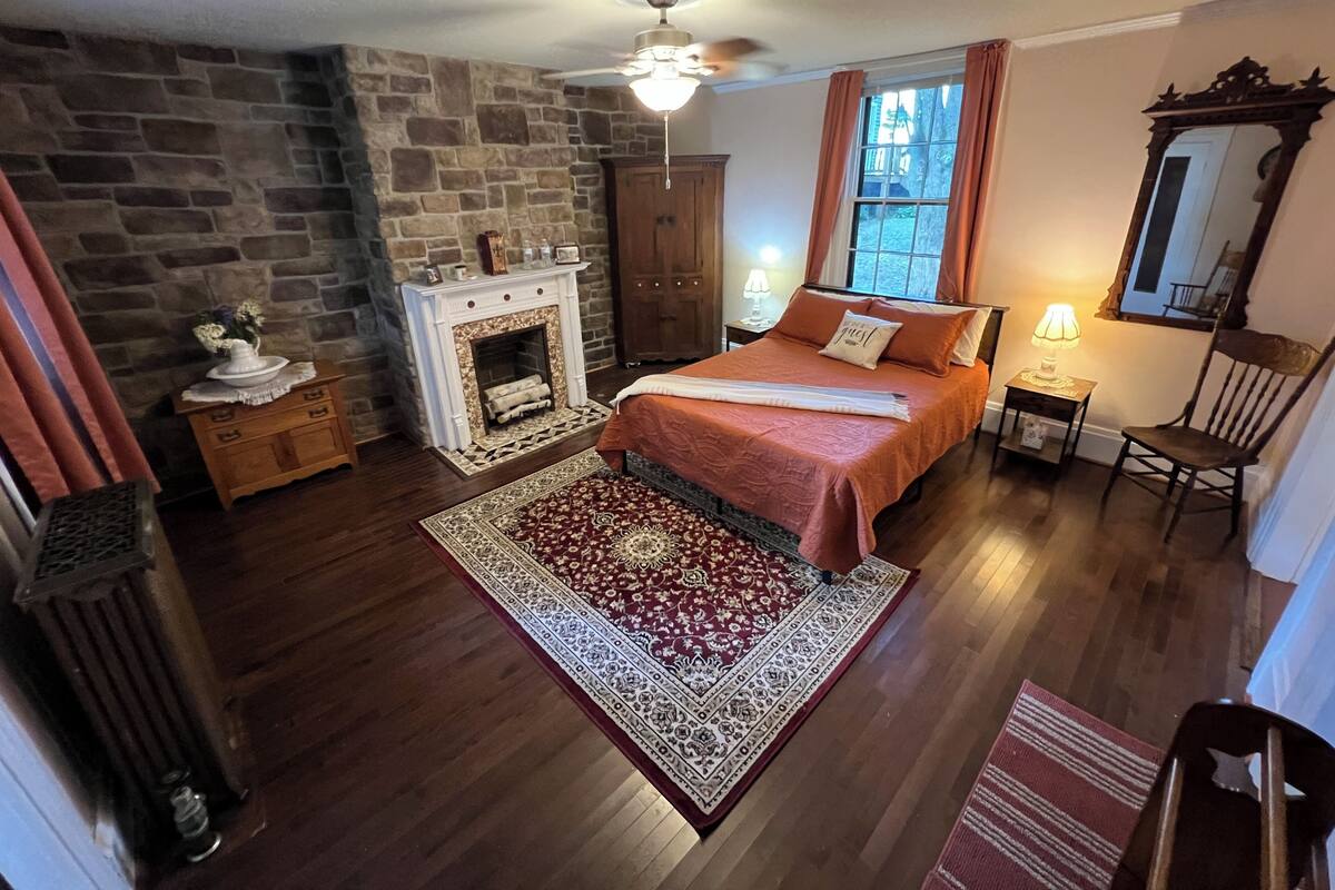 A bedroom with a stone fireplace, a bed with a red bedspread, a wooden chair, and a large mirror.