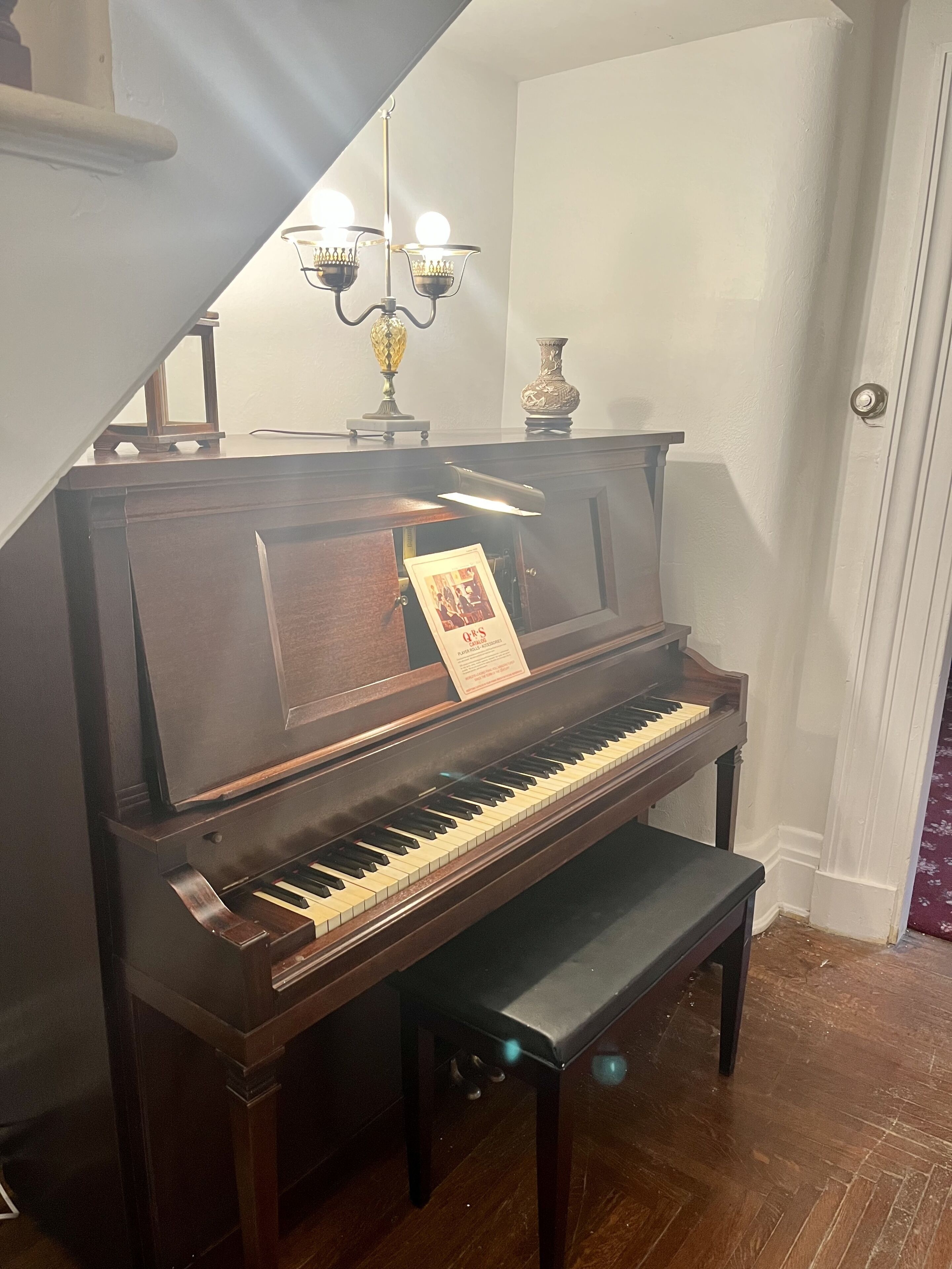 Un piano à queue dans une pièce dotée d’un plancher en bois, d’un banc, d’un lustre et d’une porte.