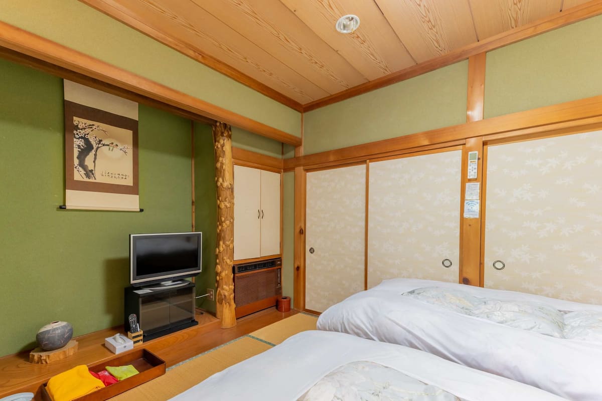 Ett hotellrum med en säng, en tv, ett träskåp och ett traditionellt japanskt konstverk på väggen.