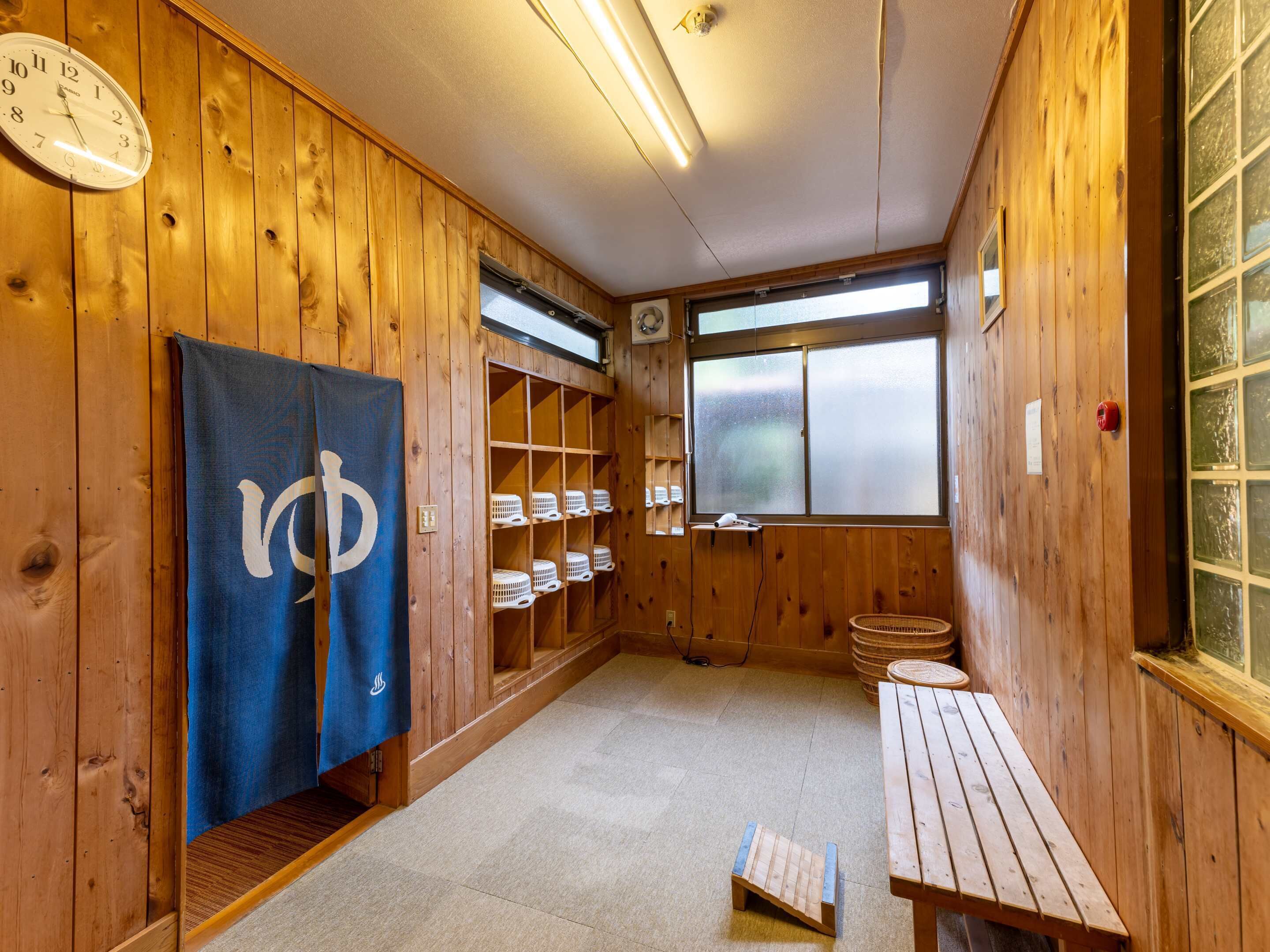 Interior de madera con un reloj, un perchero, estantes con cestas, una ventana y una pequeña mesa.