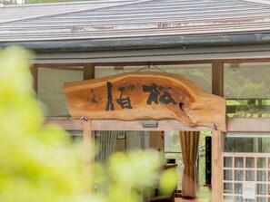 A wooden sign with Japanese characters hanging in front of a building with large windows. - hinokiyunoyado syouhaku (Azumino)