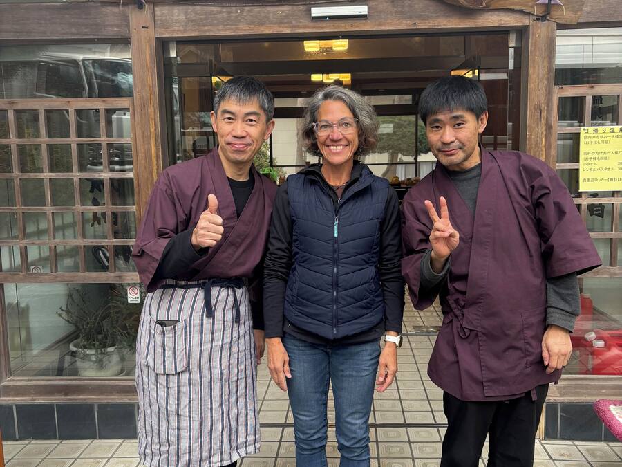 Three people standing in front of a wooden sign with Japanese characters, a tiled floor, and a glass door.