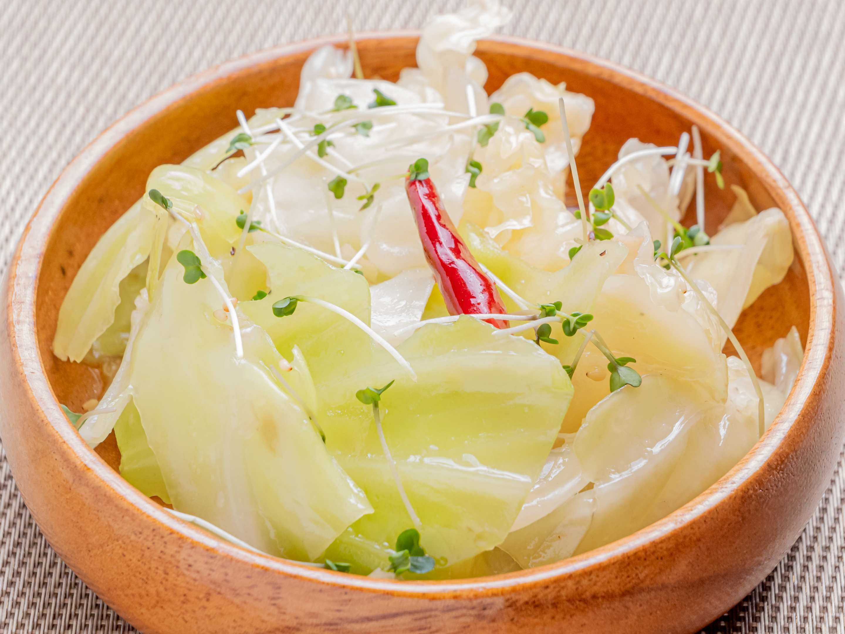 A wooden bowl filled with translucent vegetables, possibly daikon radish, garnished with sprouts and a red chili.