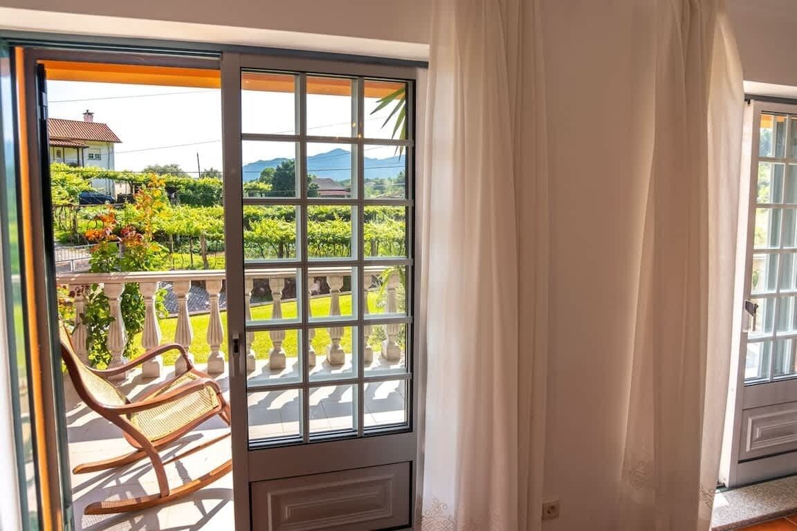 A room with a sliding glass door leading to a balcony with a view of a vineyard and a house.