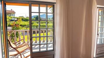 A room with a sliding glass door leading to a balcony with a view of a vineyard and a house.
