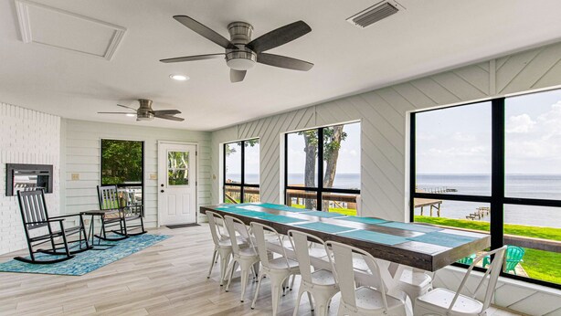 A spacious dining area with a long table and white chairs, large windows with a view of the sea, and ceiling fans.