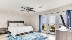 A bedroom with a bed, a ceiling fan, a large window with curtains, and a view of the outdoors.