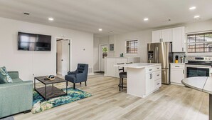 A modern kitchen with white cabinets, a stainless steel refrigerator, and a built-in oven. There is a dining area with a white table and bar stools. The living room features a green sofa and a coffee table. - Gulf Breeze Bayfront Bungalow - Dock - Pet-Friendly - Near Pensacola Beach (Gulf Breeze)