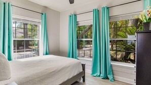 A bedroom with a bed, two windows with teal curtains, a ceiling fan, and a view of greenery outside. - Gulf Breeze Bayfront Bungalow - Dock - Pet-Friendly - Near Pensacola Beach (Gulf Breeze)