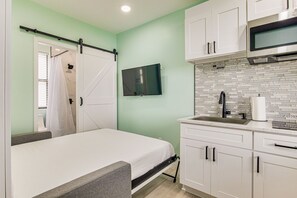A compact bedroom with a bed, a sliding door to a bathroom, a mounted TV, and a kitchenette with a microwave and sink. - Gulf Breeze Studio • Bayfront Hideaway • Sleeps 2 (Gulf Breeze)