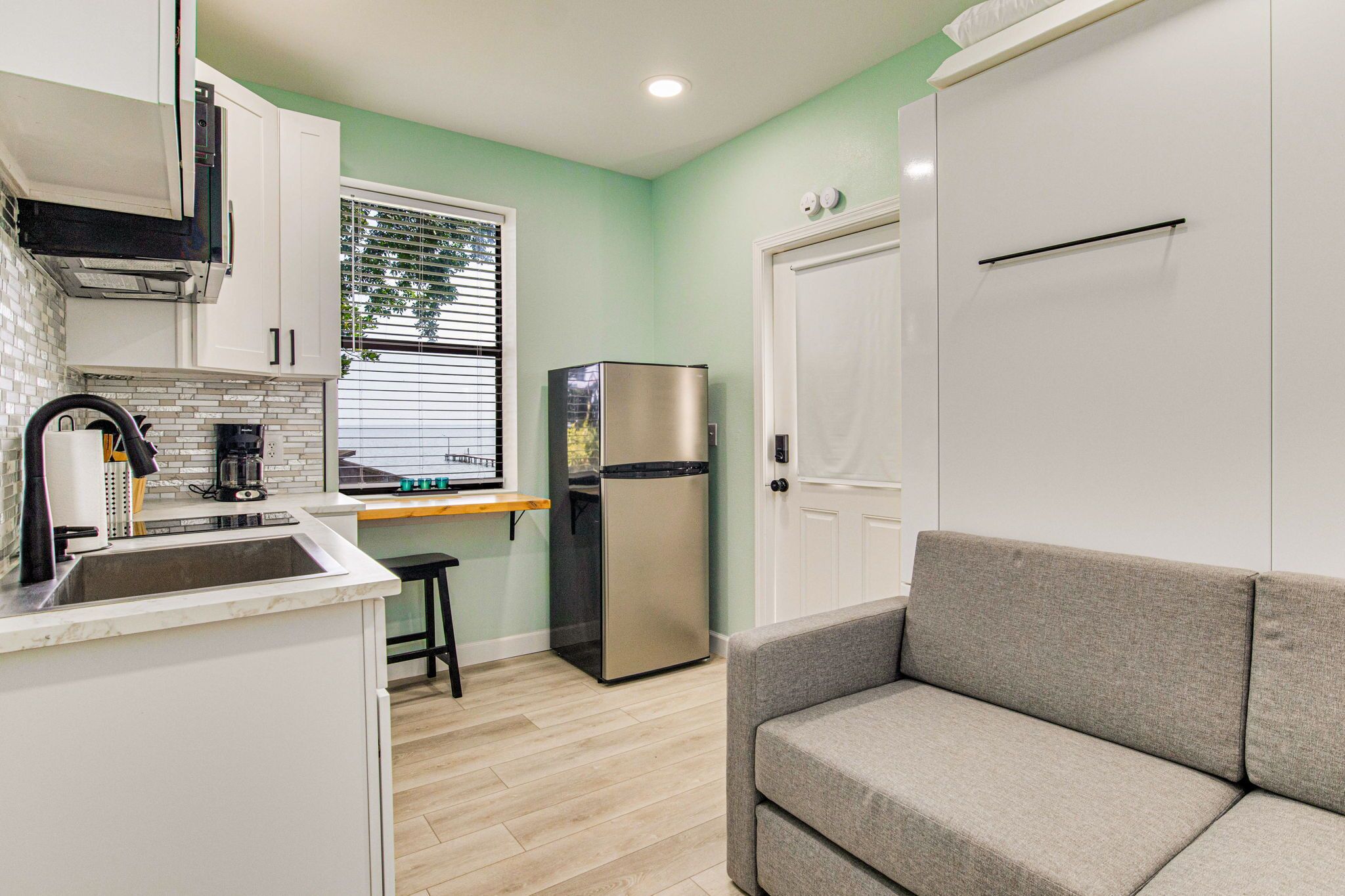 A compact kitchen with white cabinets, a stainless steel refrigerator, a coffee maker, and a window with blinds.