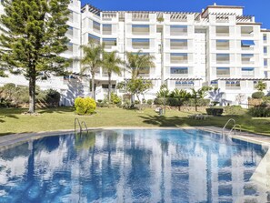 A swimming pool with a clear blue water surface, surrounded by well-maintained grass and various trees. In the background, there is a multi-story building with balconies and a white facade.