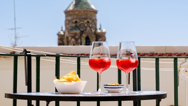 A table with two glasses of red drink, a bowl of chips, and a small dish of nuts on a balcony with a view of a historic building.