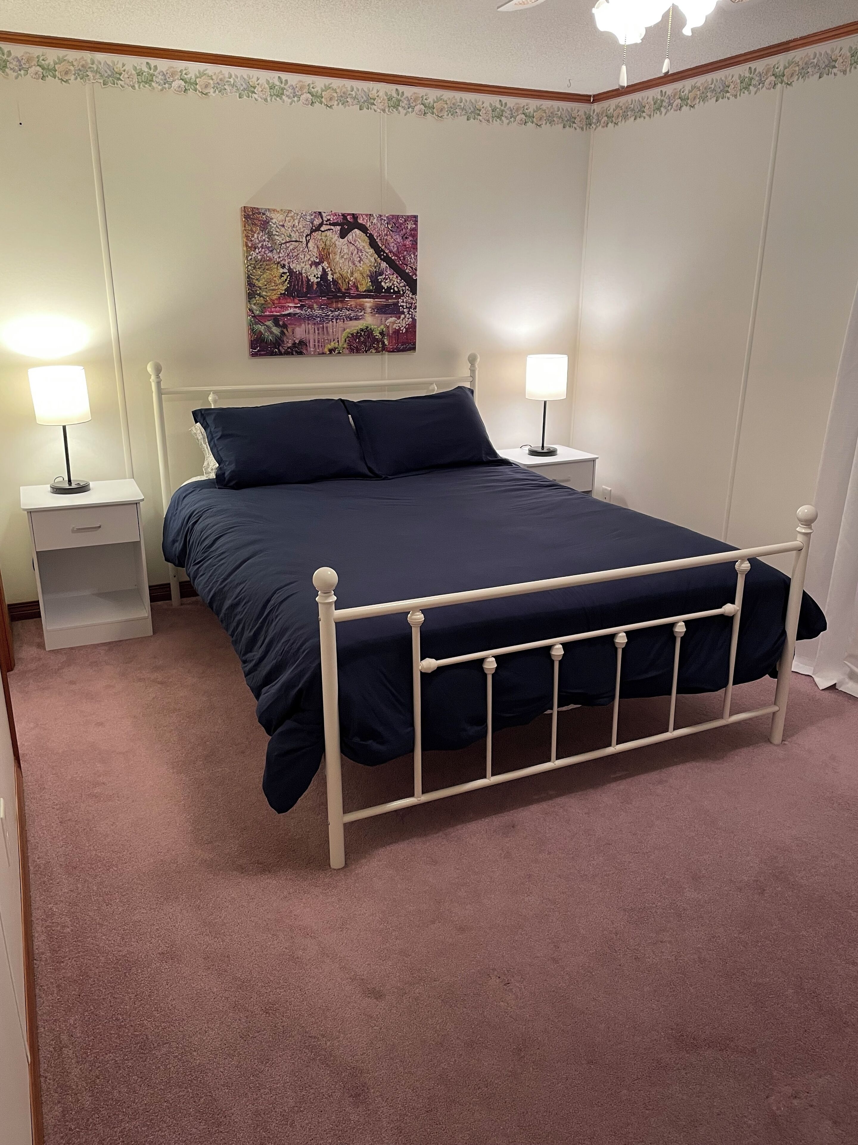 A neatly made bed with a white metal frame, two bedside tables with lamps, a floral-patterned wall, and a cityscape painting above the bed.