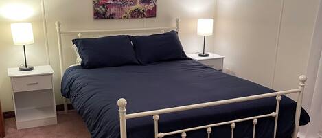 A neatly made bed with a white metal frame, two bedside tables with lamps, a floral-patterned wall, and a cityscape painting above the bed.