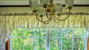 A dining area with a wooden table and chairs, a chandelier, and a view of a green landscape through a window with floral curtains. - Cozy two bedroom home with ocean view. Quiet and secluded location.  (Lubec)