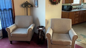 A living room with two armchairs, a small table, a wall-mounted lamp, a framed picture, and a kitchen area in the background. - Cozy two bedroom home with ocean view. Quiet and secluded location.  (Lubec)