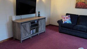 A living room with a flat-screen TV, wooden entertainment unit, red carpet, grey sofa, and a ceiling fan. - Cozy two bedroom home with ocean view. Quiet and secluded location.  (Lubec)