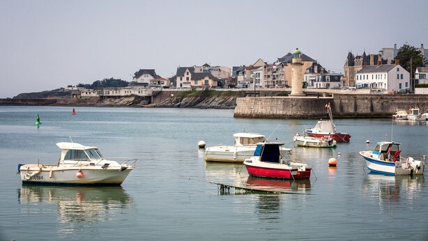 A harbor with several boats docked, a stone quay, and buildings in the background.