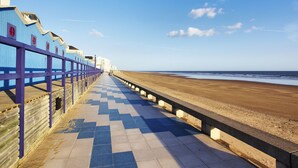 A seaside promenade with a checkered walkway, wooden buildings with purple railings, and a sandy beach.