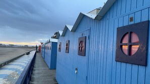 A row of blue wooden buildings with windows, a wooden walkway, and a view of the beach.