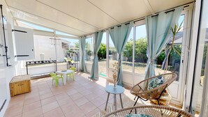 Un patio iluminado por el sol con una mesa y sillas de comedor, una silla de mimbre y una estantería.