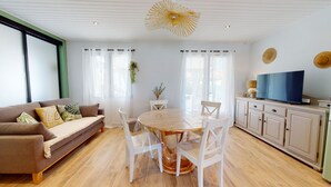 Una sala de estar moderna con una mesa de comedor de madera, sillas blancas, un sofá color beige y un televisor sobre un mueble.