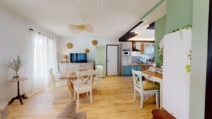Una cocina acogedora y bien iluminada con armarios de madera, un comedor con mesa y sillas, y un espacio de sala de estar con televisor y objetos decorativos.