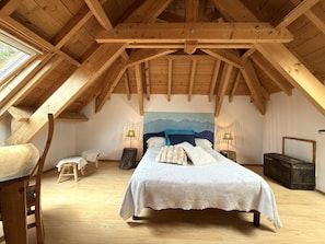 Une chambre avec un plafond en bois, un lit avec une literie blanche, un tableau représentant des montagnes et un petit tabouret.