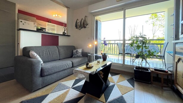 Una sala de estar moderna con un sofá gris, una mesita de centro y un tapete estampado. Hay un ventanal corredero que da a un área exterior con balcón.