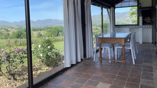 A covered patio with a table and chairs, large windows offering a view of a green landscape with hills.