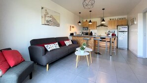 Una sala de estar moderna con un sofá gris, cojines rojos, una pequeña mesa blanca y una cocina con gabinetes de madera y un refrigerador de acero inoxidable.