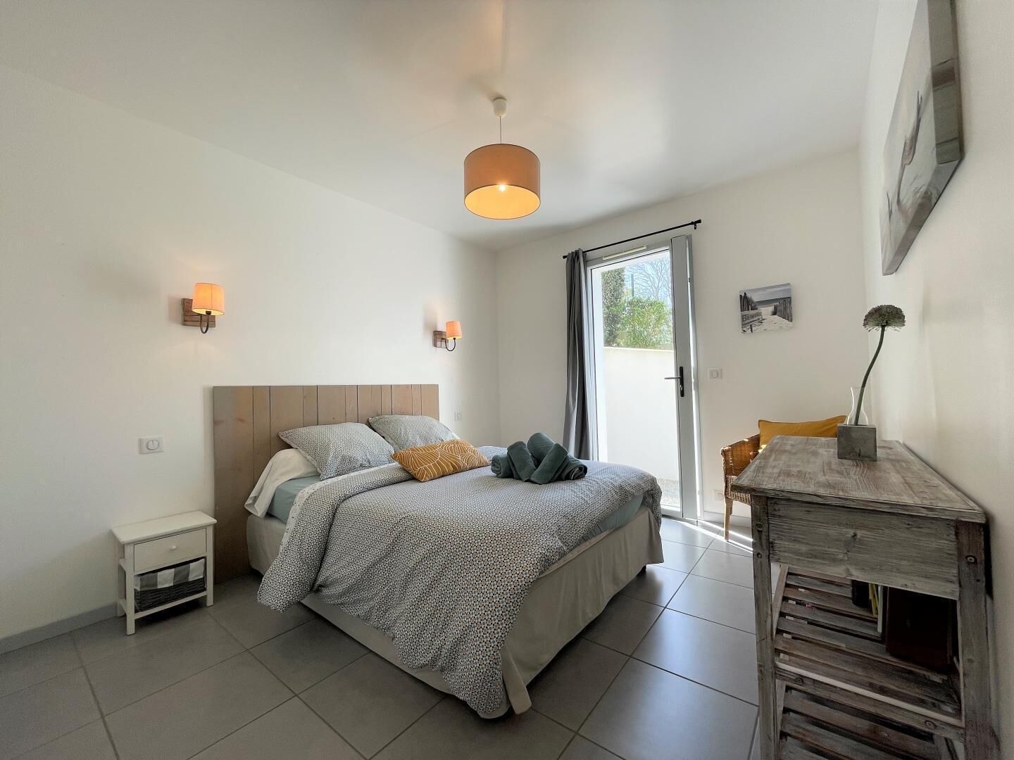 A bedroom with a bed, bedside tables, a nightstand, a wooden side table, and a view through a glass door.