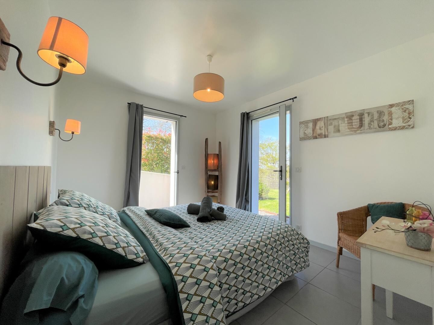 Een slaapkamer met een bed, twee wandlampen, een schuifdeur met glas, een rieten stoel en een tafeltje met een vaas.
