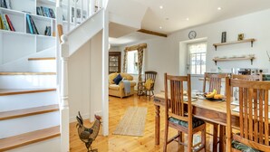 Un vaste espace de vie agrémenté d’un escalier en bois, d’une table à manger avec des chaises, d’une bibliothèque et d’un coq décoratif.