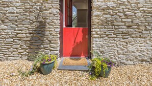 Un bâtiment en pierre avec une porte rouge, deux jardinières suspendues et des plantes en pot disposées au sol.