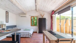 Quarto compacto com cama, uma mesinha, um banco, janela e porta que dá para uma área externa.