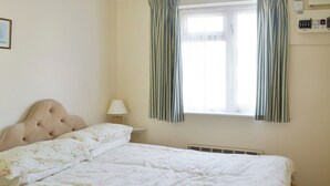 A bedroom with a large bed, a nightstand, a lamp, a window with curtains, and a wall-mounted electrical panel. - Mariners Rest - UK44498 (Hope Cove, near Kingsbridge)