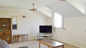 A living room with a TV, wooden furniture, a chandelier, and a window with curtains. - Mariners Rest - UK44498 (Hope Cove, near Kingsbridge)