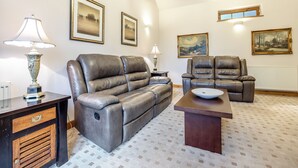 Un salon comprenant un canapé en cuir, un fauteuil en cuir, une table basse en bois et une table d’appoint avec une lampe. Les murs sont ornés de tableaux encadrés et un lustre est suspendu au plafond.