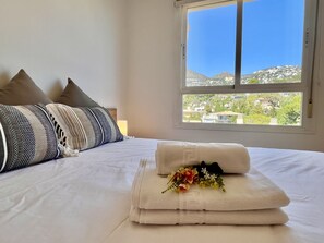 A hotel room with a bed, pillows, a window with a view of buildings and hills, and a bouquet of flowers on the bed.