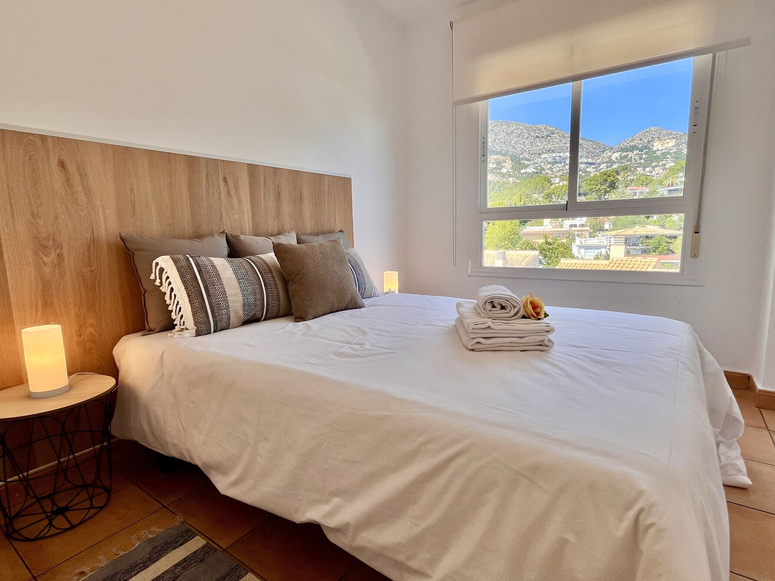 Ein Schlafzimmer mit einem Bett, einem Nachttisch, einer Lampe und einem Fenster mit Blick auf einen Berg und Gebäude.