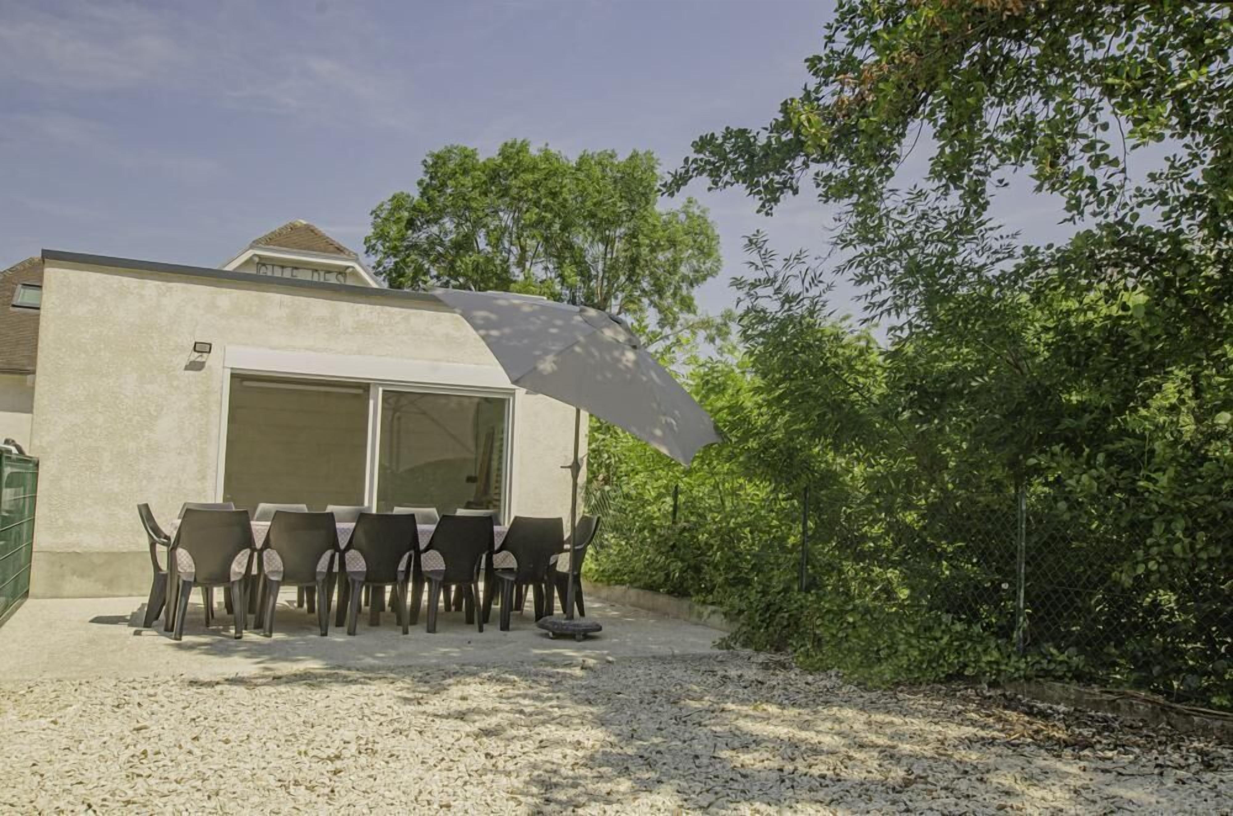 Un espace extérieur aménagé pour les repas, avec une table et des chaises, un coin salon abrité et, au fond, un bâtiment avec une porte de garage.